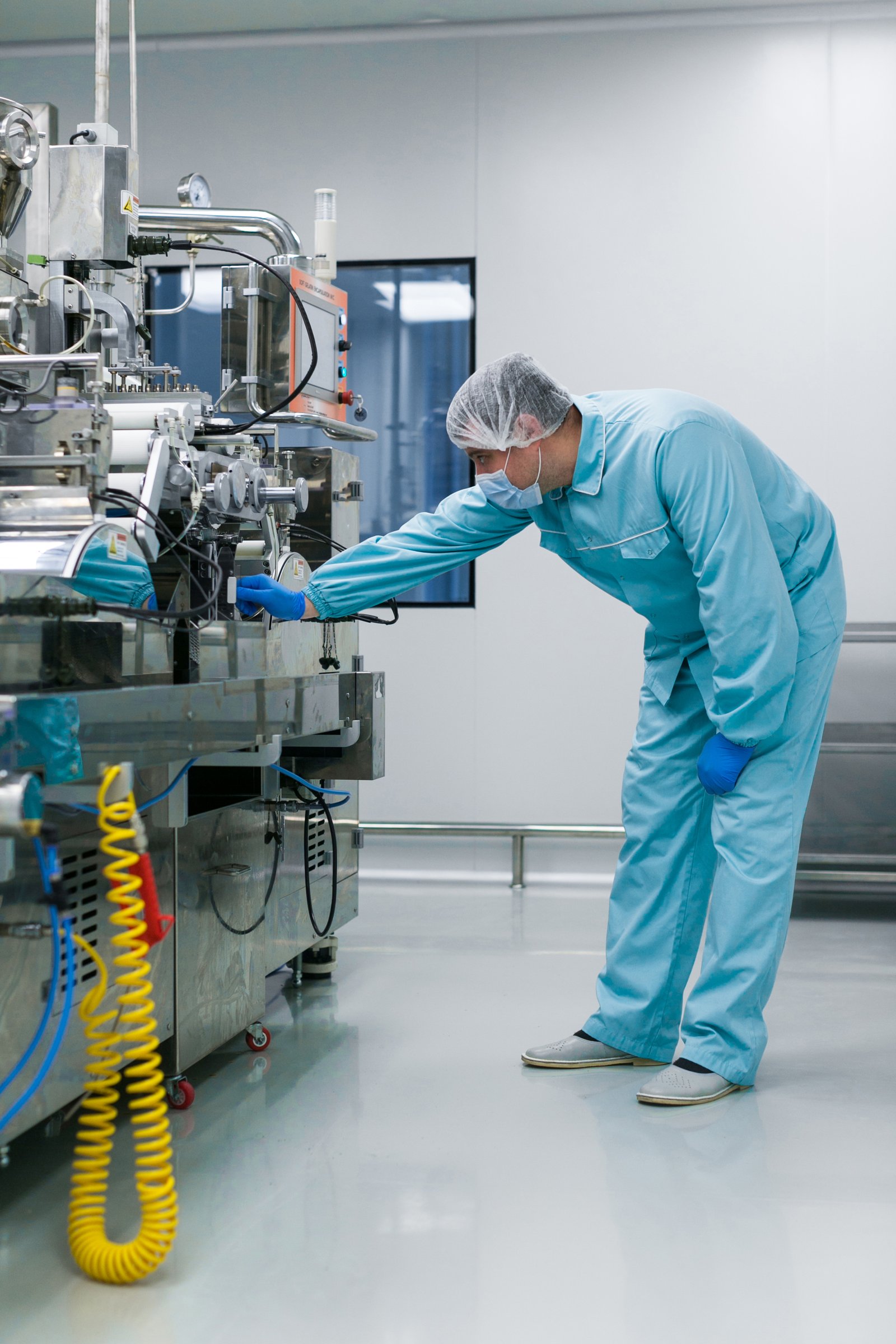 vertical picture of bended caucasian scientist in blue lab suit trying to fix manufacture machine with shafts in clean factory